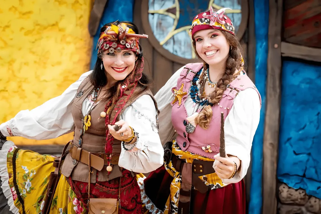Dos mujeres sonrientes con coloridos trajes de estilo gitano, pañuelos en la cabeza y collares posan delante de una fachada de madera de colores vivos