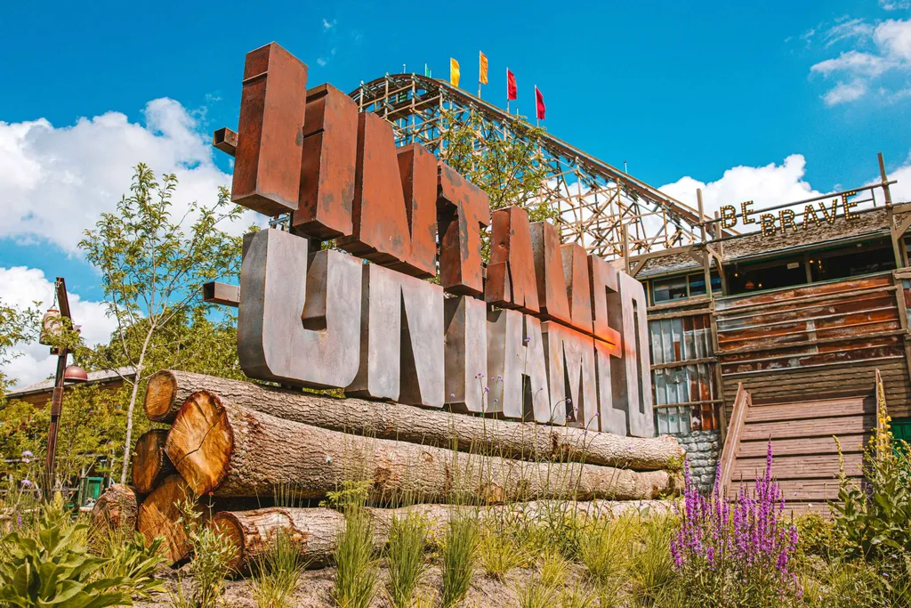 Grandes letras oxidadas del cartel de Untamed sobre troncos, con una montaña rusa de madera y un edificio con 'Be brave' detrás, en Walibi Holland
