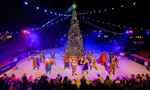 Artistas con trajes de invierno bailan de noche en una pista de hielo en Toverland, ante un árbol de Navidad iluminado y el público mirando Artistas con trajes de invierno bailan de noche en una pista de hielo en Toverland, ante un árbol de Navidad iluminado y el público mirando