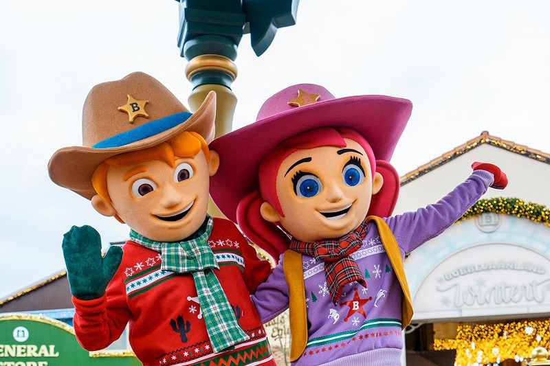 Dos personajes disfrazados con atuendo de vaquero y jerséis navideños saludan frente a un edificio con decoración invernal y el rótulo Bobbejaanland Wintert
